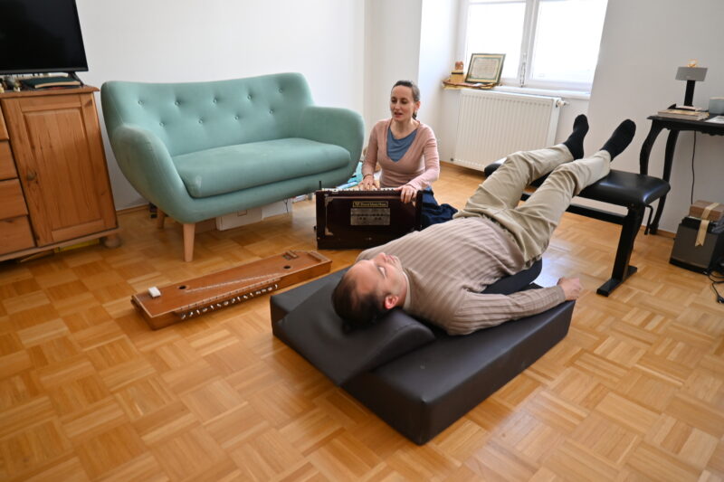Indische Musiktherapie und Musik - Energetik mit Schumann 3D Platte. Elisabeth Schulz sitzt am Boden und singt und spielt Harmonium. Der Kunde liegt aud der 3D Platte